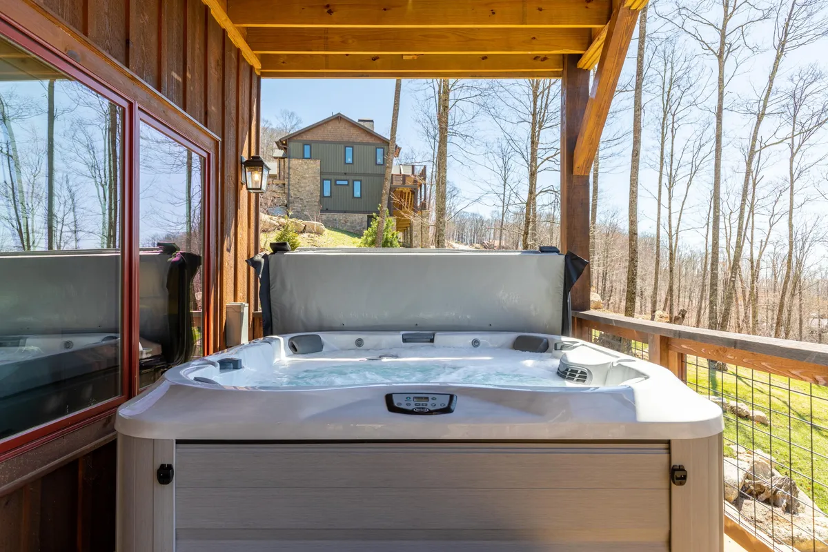 An inviting outdoor hot tub sits on a wooden deck, surrounded by lush trees and distant glimpses of another house. The setting offers privacy and a direct connection to the beautiful natural surroundings. The scene suggests tranquil soaking while gazing out into the woods or up at the stars.