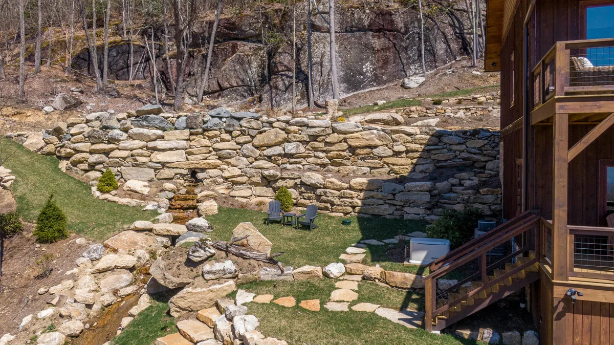 A tranquil outdoor seating area nestled beside a stone wall and a gently flowing stream. The scene includes a pathway leading to a restful spot, ideal for quiet reflection, morning coffee, or relaxed socializing surrounded by natural beauty.