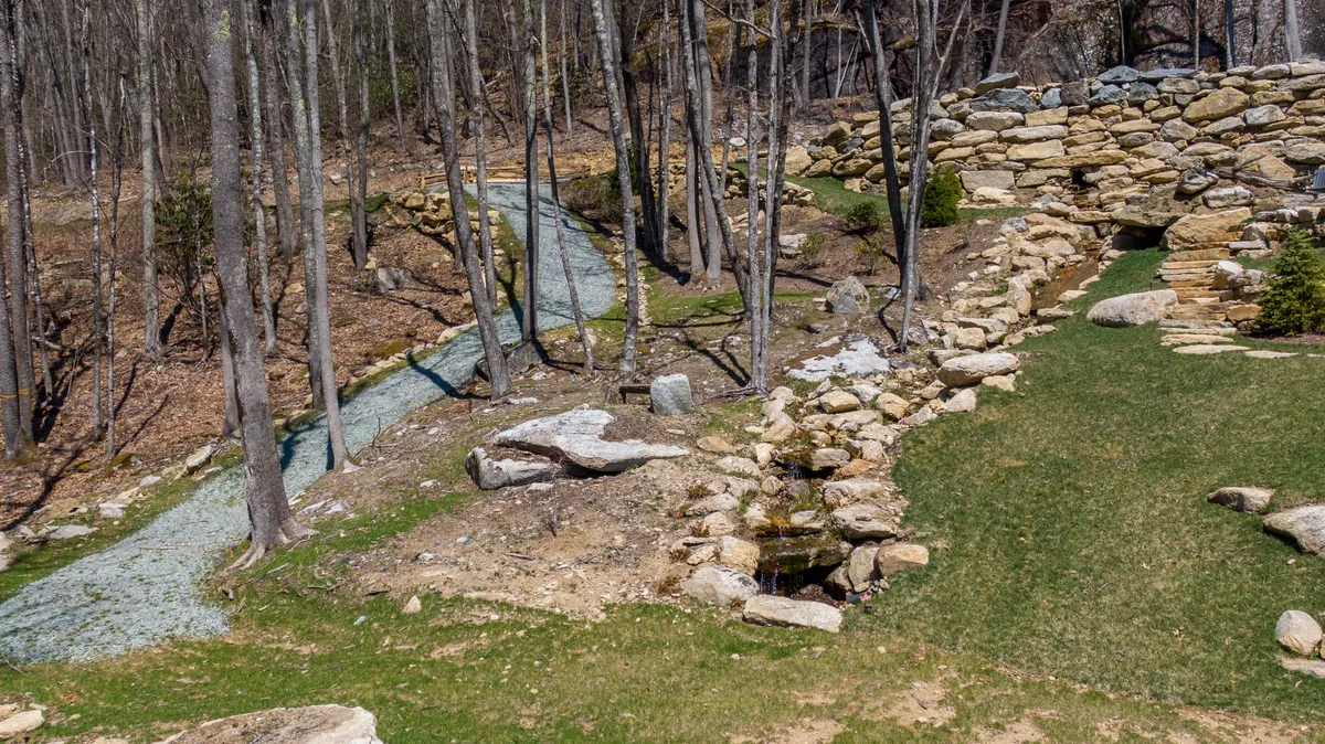 A peaceful exterior scene with rock formations, a meandering path, a water feature, and abundant trees. The landscape naturally encourages exploration, relaxation, and interaction with nature, suggesting a variety of outdoor activities.