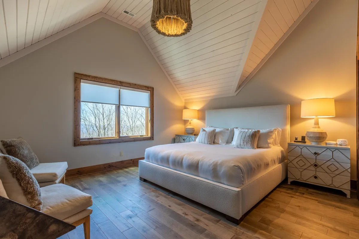 A spacious king bedroom with a large, comfortable bed, a decorative light fixture above, nightstands on either side, and a window providing views and natural light. The setting is serene, emphasizing relaxation and the comforts of a restful retreat.