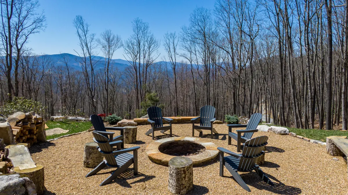 This exterior image features a fire pit with Adirondack chairs arranged for group seating, set against a backdrop of a sweeping mountain view and bare trees hinting at seasonal change. The scene evokes a sense of camaraderie and enjoyment of the outdoors, with open skies perfect for stargazing and mountain sunsets.