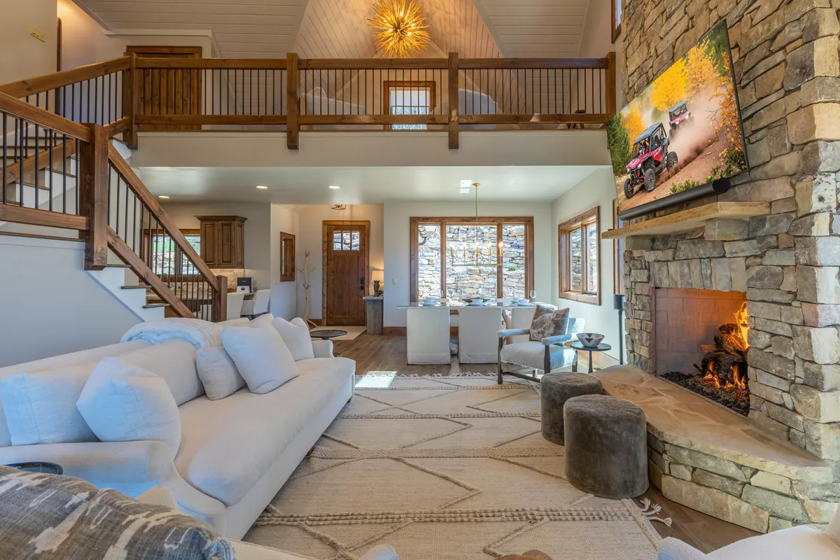 A spacious open-concept living room featuring a large sectional sofa arranged around a prominent stone fireplace. High ceilings add to the airy atmosphere, while a wooden staircase and a TV mounted on the stone wall complete the setting. Ample natural light spills in, enhancing the sense of warmth and inviting togetherness.