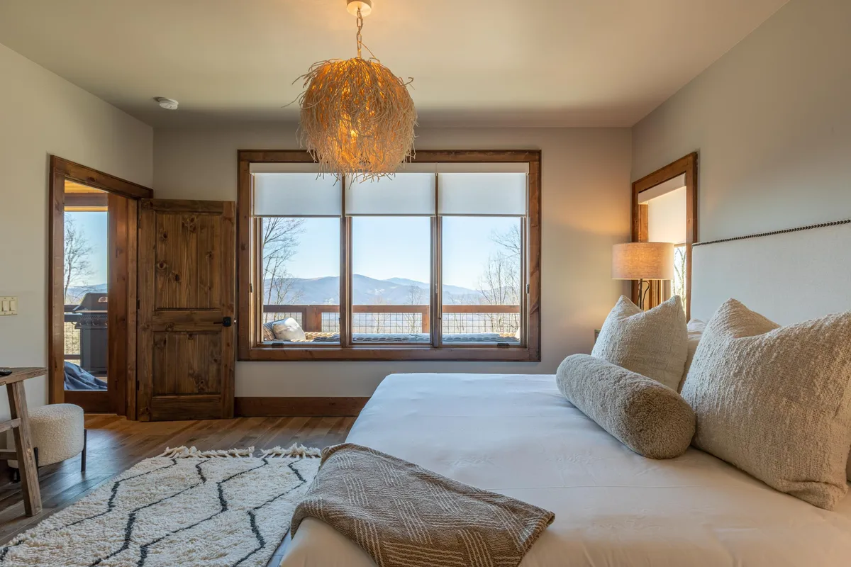 A bright and inviting king bedroom with a large bed featuring decorative pillows, stylish lighting, and expansive windows that frame picturesque mountain views. The space exudes tranquility and comfort, perfect for peaceful mornings or restful evenings.