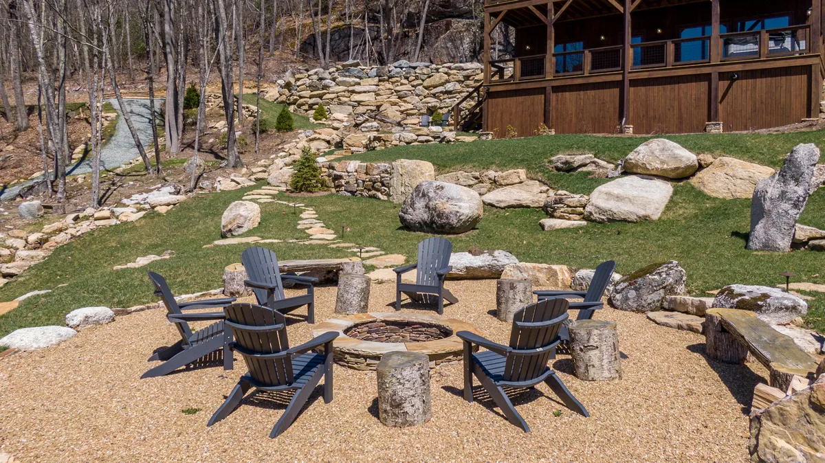This scene spotlights a fire pit surrounded by Adirondack chairs and a stone pathway, with the cabin structure and carefully landscaped greenery in the background. The arrangement suggests an inviting space for small group gatherings, evening chats, and enjoying the peaceful mountain air.
