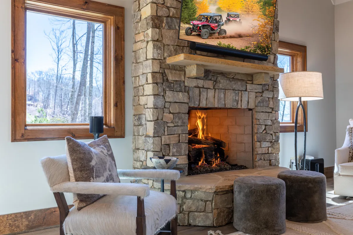 This interior scene displays a cozy lounge area featuring a stone fireplace, a large television, comfortable sofas arranged for easy conversation, and expansive windows that let in natural light and offer exterior views. The space communicates warmth and is clearly designed for both relaxation and social gatherings, with the TV and seating arrangement inviting movie nights or group entertainment.