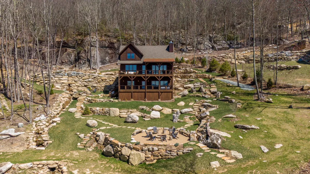The image features the exterior of a luxurious mountain home set amidst natural rock formations and lush landscaping. A stone pathway leads to a welcoming fire pit area, perfect for gathering, with the house overlooking expansive sunset views from its balcony. The surrounding scenery evokes tranquility, inviting guests to relax outdoors and soak in the beauty of Banner Elk.