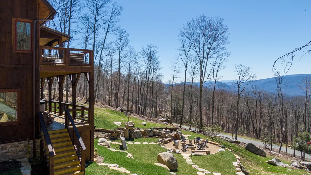 A spacious wooden deck with a stone pathway leading to a cozy fire pit area, surrounded by trees and mountain views in the distance. The space is ideal for gathering after a day outdoors, relaxing, or stargazing under clear skies.