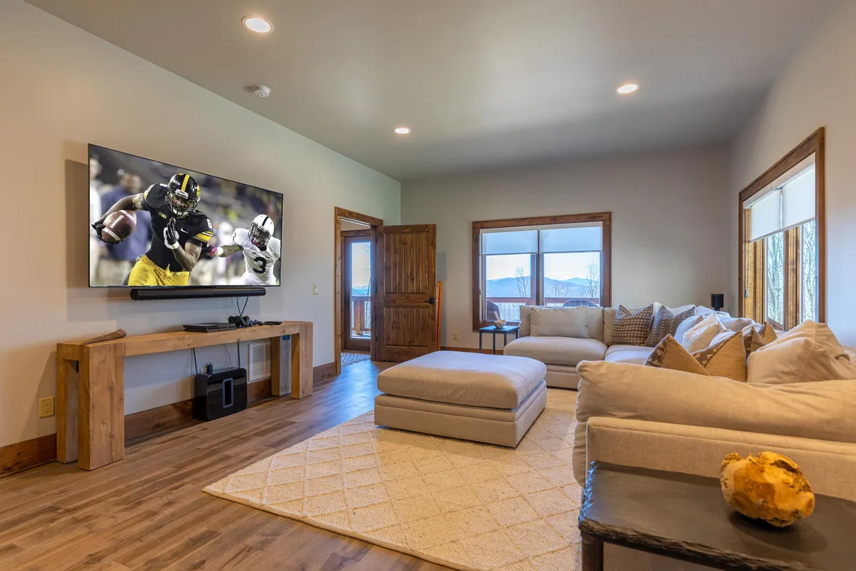 The image showcases a spacious sectional sofa facing a large wall-mounted television with a wooden coffee table in front. Natural light pours in from nearby windows, creating a bright, welcoming ambiance. The layout is ideal for both lively group gatherings and quiet solo relaxation, with ample room for everyone to spread out.